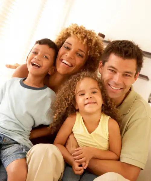 A smiling family of four sits closely together indoors, with two adults and two young children, all looking happy and relaxed—enjoying peace of mind that comes with life insurance in Orange County, CA.