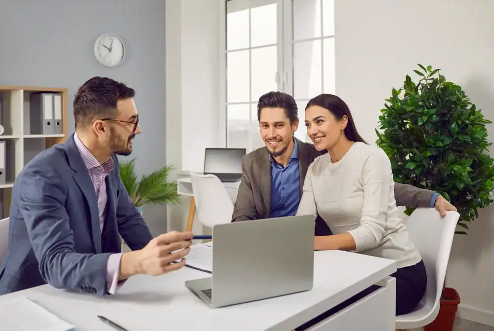 A man in a suit talks to a smiling couple seated across from him at a desk with a laptop in a bright office, suggesting a professional consultation about life insurance Orange County or auto insurance Orange County.