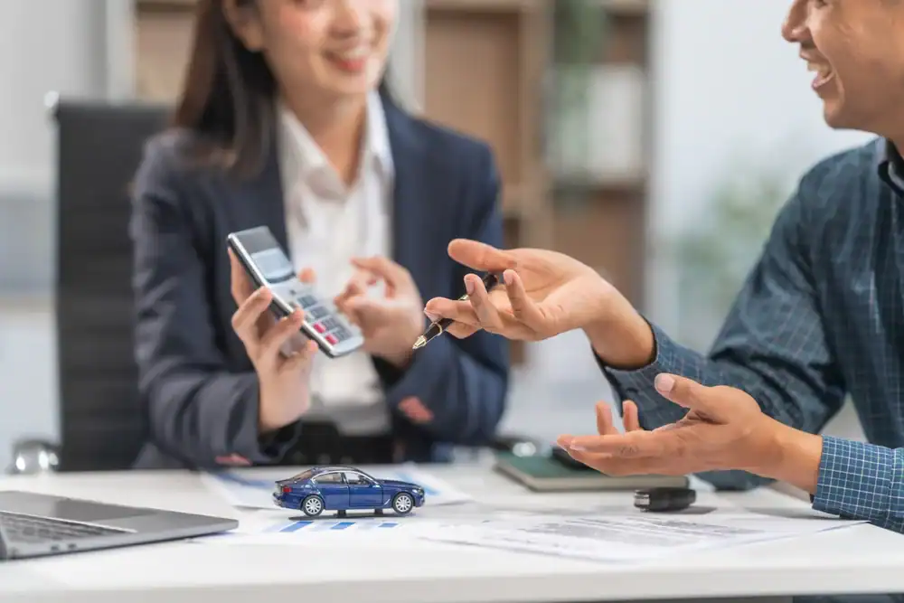 A person hands over car keys to another across a desk with documents, a model car, and a laptop&mdash;both smiling, discussing auto insurance Orange County, CA as one holds a calculator.