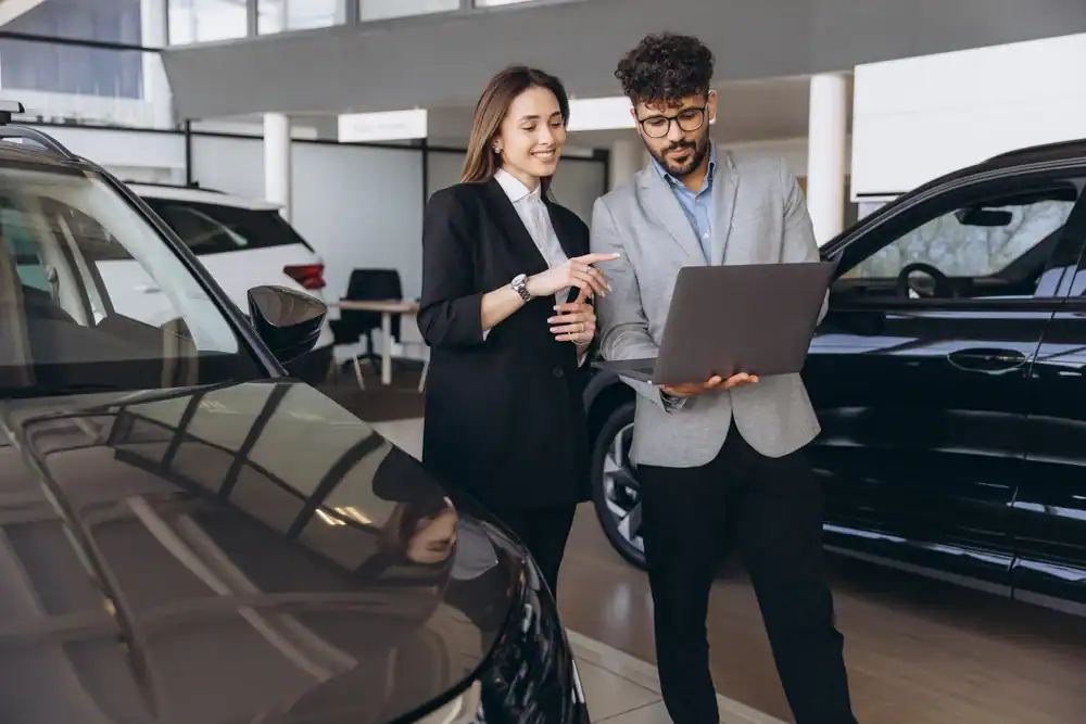 A man and woman in business attire stand between cars in a CA dealership, discussing something on a laptop, with cars visible around them in a bright showroom&mdash;perhaps reviewing auto insurance Orange County options.