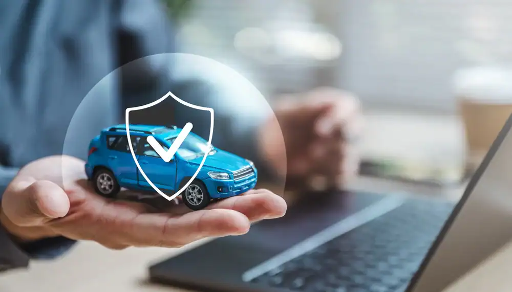 A person holds a small blue car model with a shield and checkmark graphic overlay, symbolizing auto insurance Orange County, in front of an open laptop on a desk.