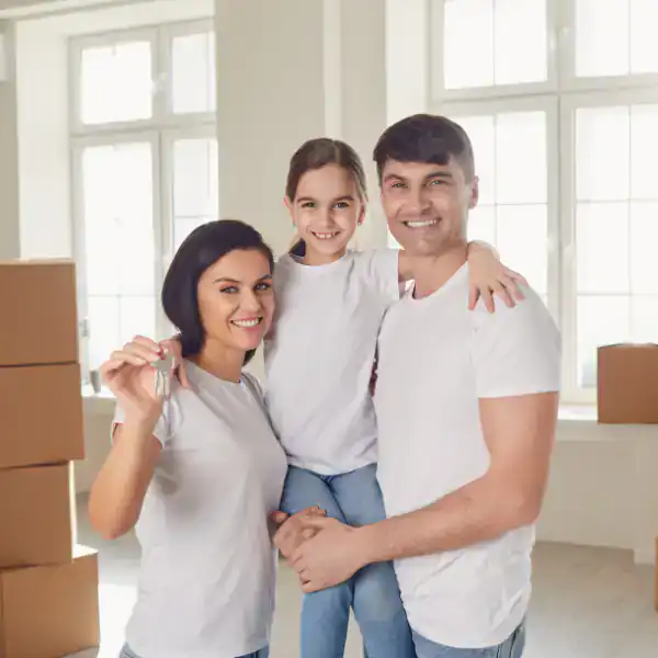 A smiling family of three, all in white shirts, stands in a bright room with cardboard boxes. The mother holds house keys, the father holds their daughter—happy and secure in their new home with life insurance Orange County coverage.