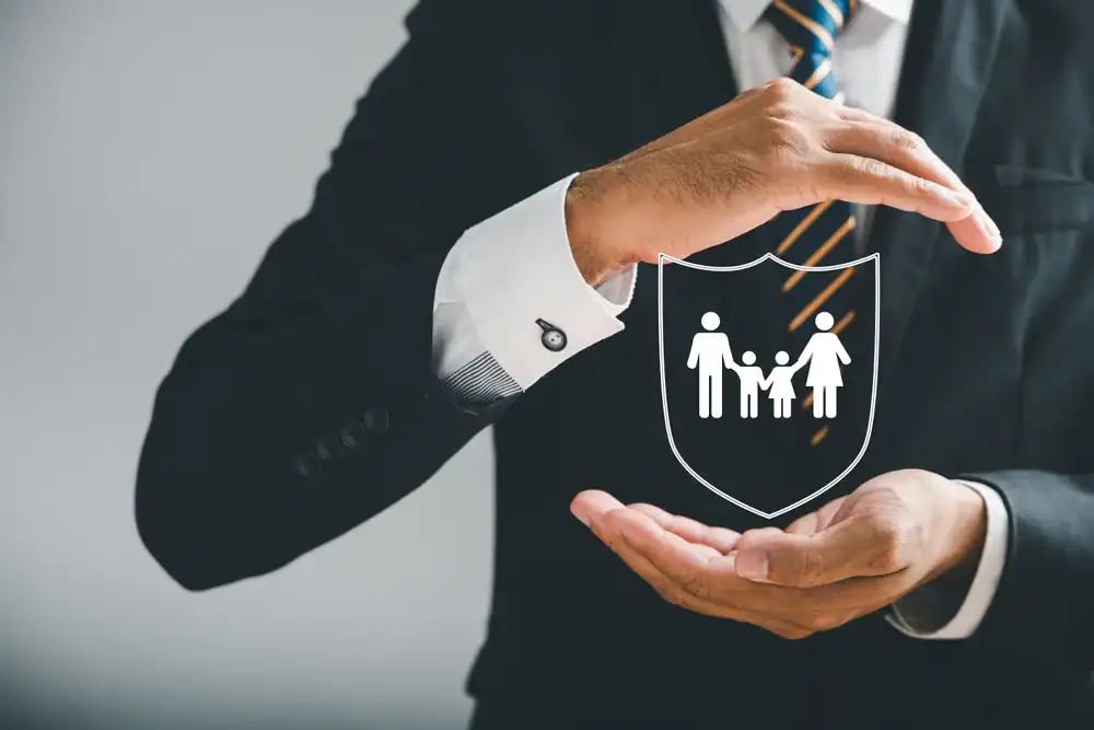 A man in a suit holds his hands protectively around a shield with a family icon, symbolizing life insurance Orange County and family protection, as well as the value of safety coverage in CA.