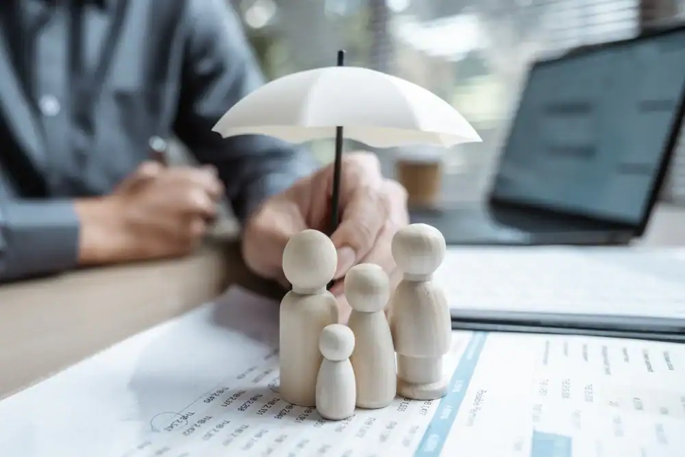 A hand holds a white umbrella over four wooden figurines representing a family, placed on financial documents&mdash;a reminder of the importance of life insurance Orange County for protecting your loved ones. A laptop and person are visible in the background.