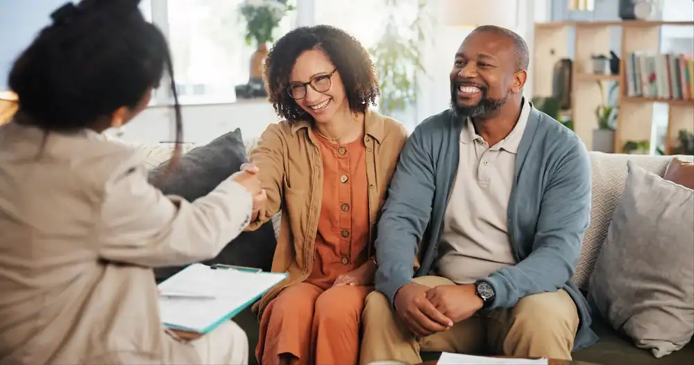 A smiling couple sits on a couch, shaking hands with a professional across from them&mdash;discussing life insurance Orange County, CA&mdash;in a cozy, well-lit office or living room with books and plants in the background.