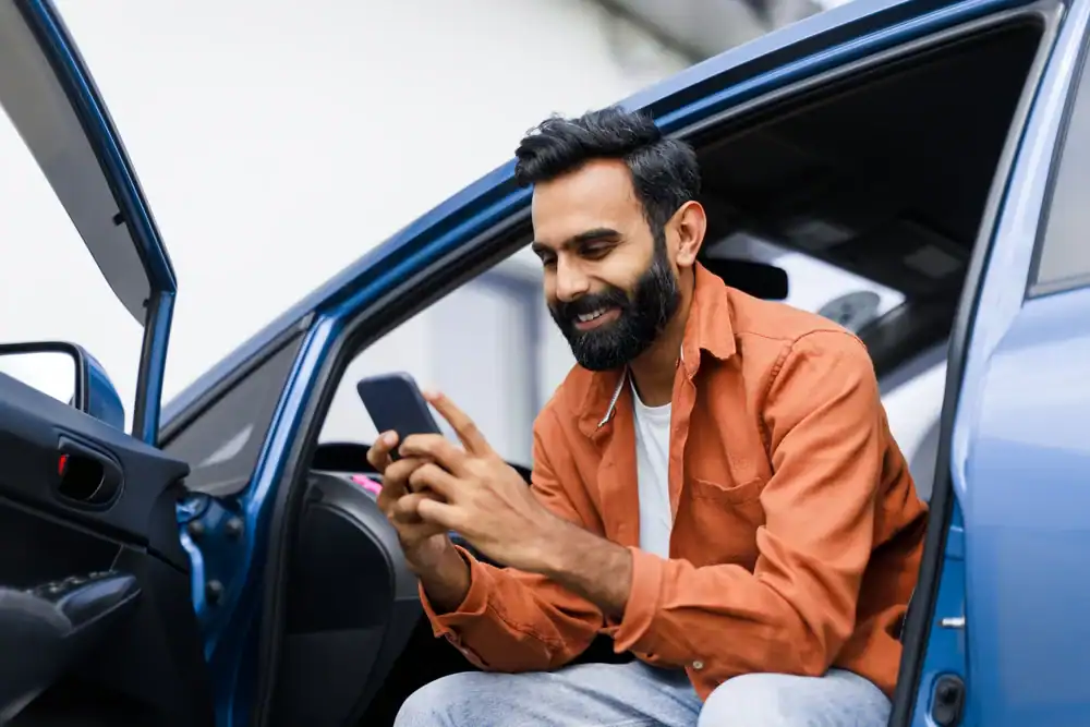 A man with a beard, wearing an orange shirt and white t-shirt, sits in the open doorway of a blue car, smiling while looking at his smartphone&mdash;perhaps checking his auto insurance Orange County, CA options.