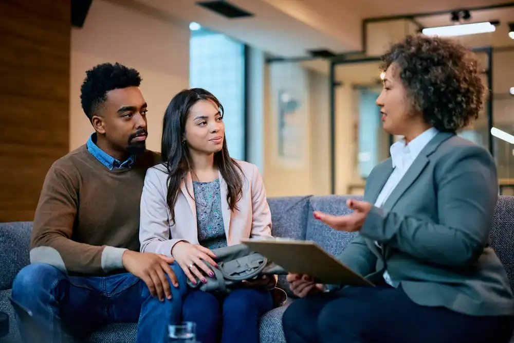 A couple sits closely together on a sofa, attentively listening to a professional woman in a suit who is holding a clipboard and discussing life insurance Orange County, CA, in a modern office setting.