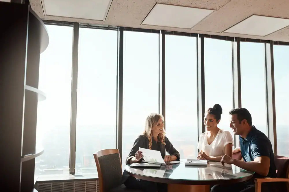 Three people sit at a round table in a modern Orange County office with large windows, discussing life insurance documents and smiling, as sunlight streams in from outside.
