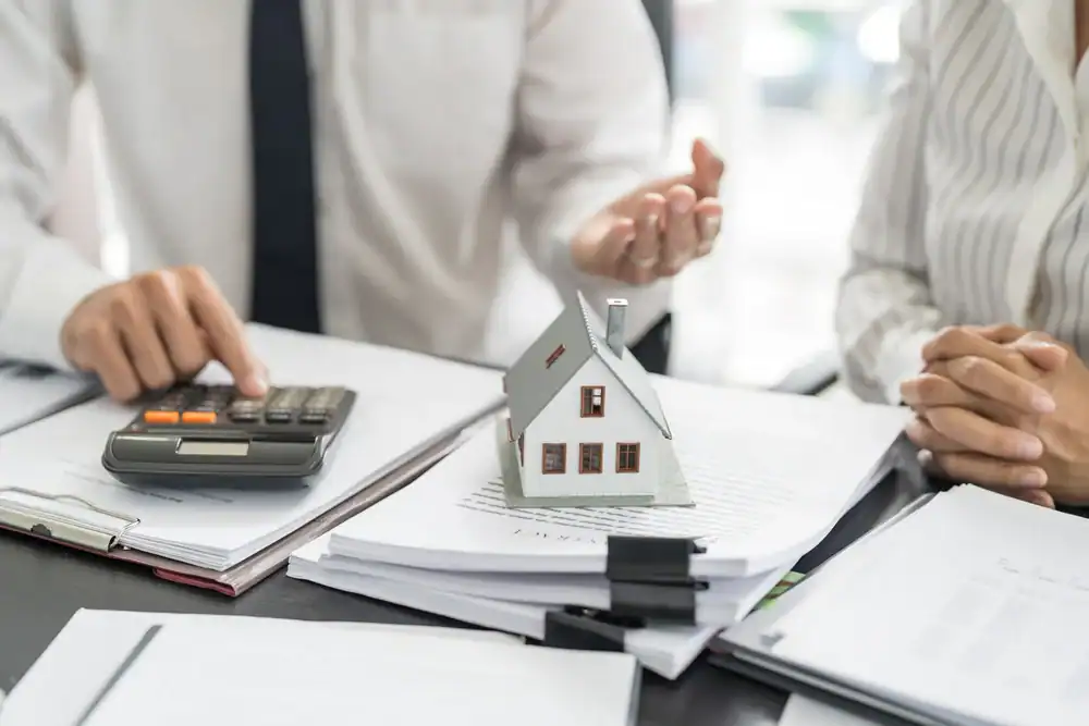 Two people sit at a desk with paperwork, one using a calculator and the other gesturing. A small model house rests on the documents, suggesting a discussion about real estate, life insurance Orange County, CA, or mortgages.