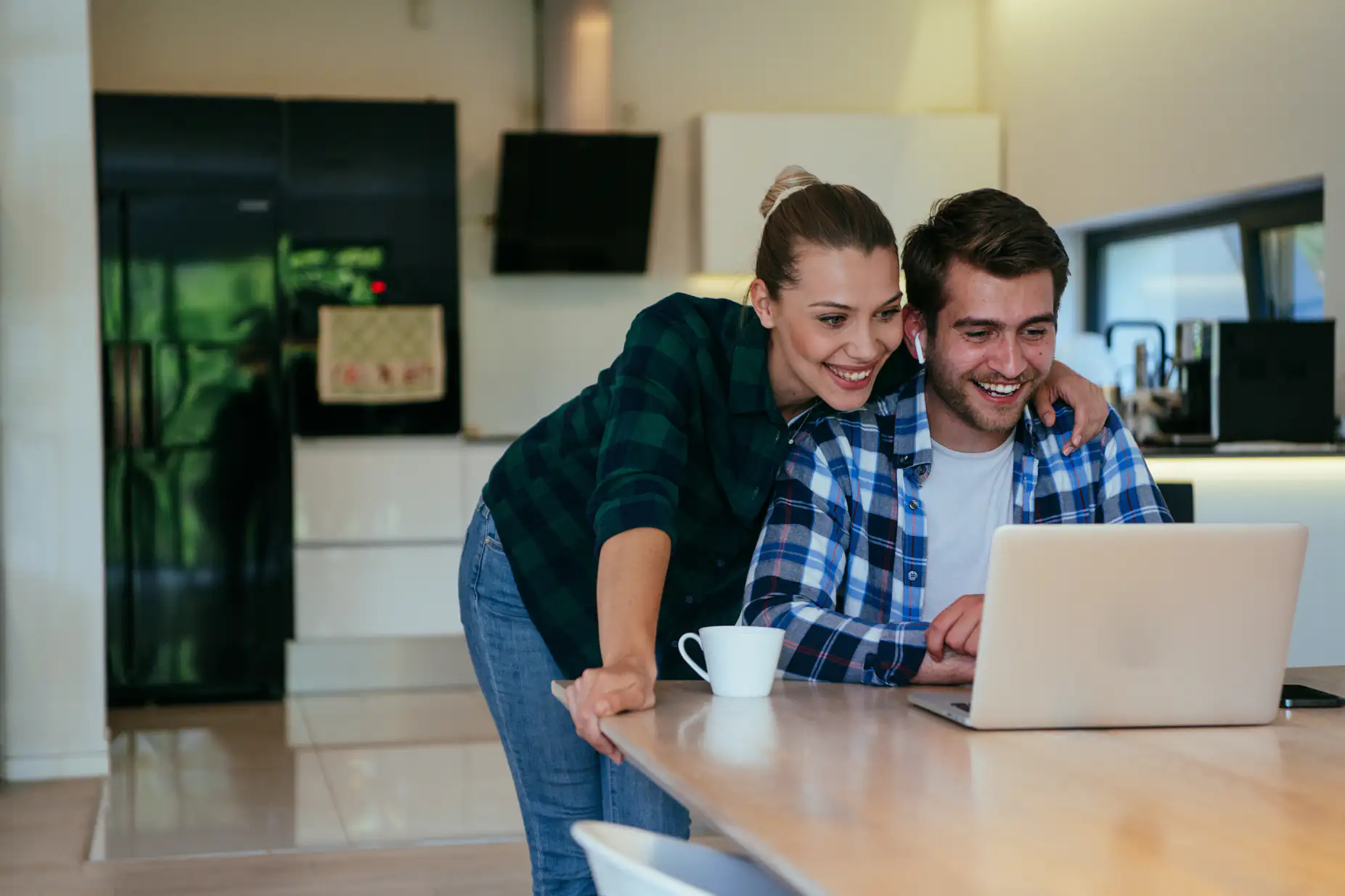 A smiling couple in casual clothes looks at a laptop together in a modern kitchen. The woman leans on the table beside a coffee cup, while the man sits and uses the laptop.