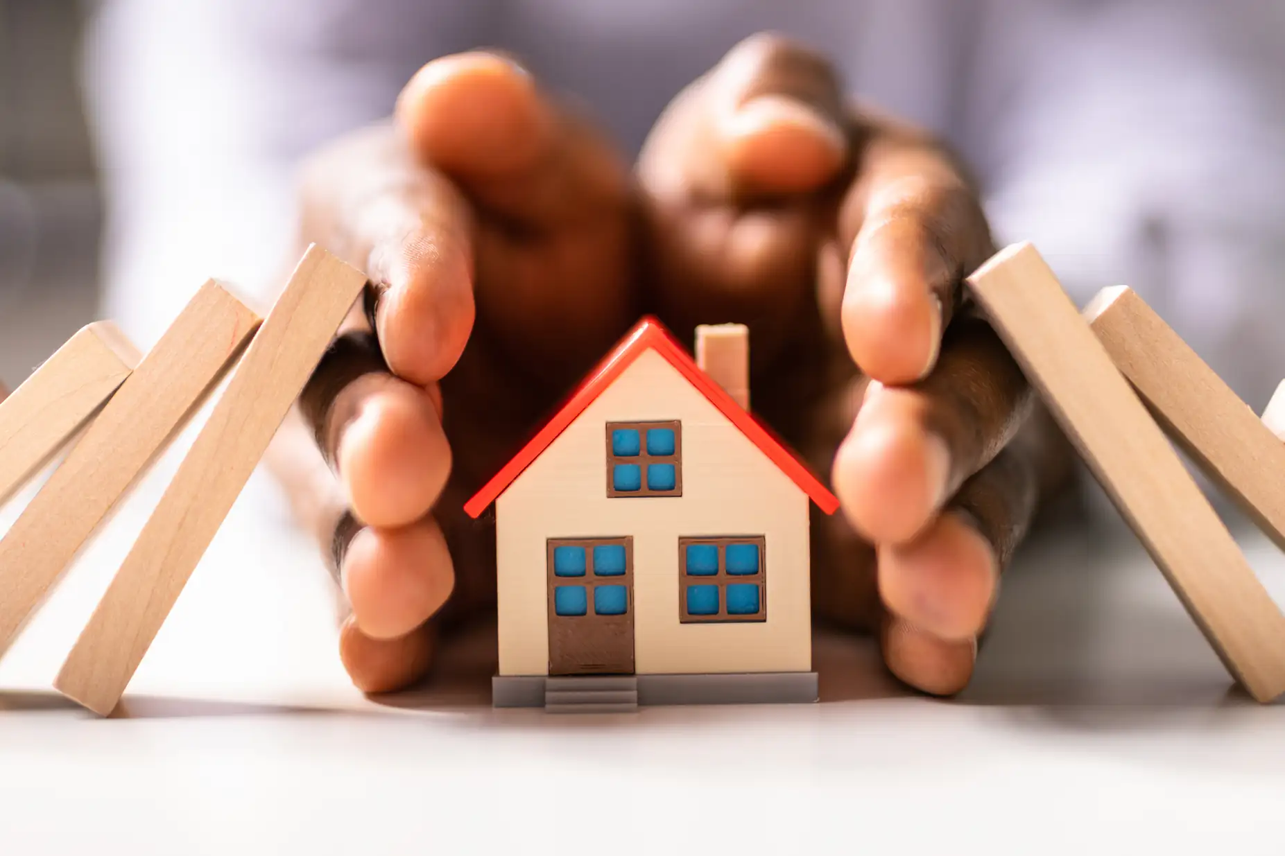 A pair of hands protectively surrounds a small model house, preventing falling wooden dominoes on each side from knocking it over, symbolizing home protection and security.